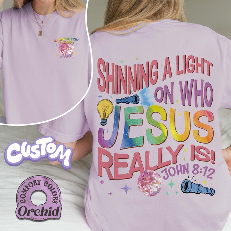 May include: Light purple t-shirt with the text "Shining a Light on Who Jesus Really Is!" in colorful letters. The shirt also features a graphic of a lightbulb, a telescope, and a disco ball. The front of the shirt has the text "Illumination Station".