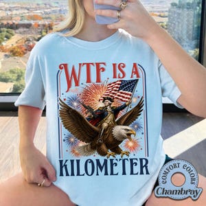 May include: Light blue t-shirt with a patriotic graphic. The design features an eagle carrying a figure holding a flag, with fireworks and the text "WTF IS A KILOMETER". The shirt has a round neckline and short sleeves.