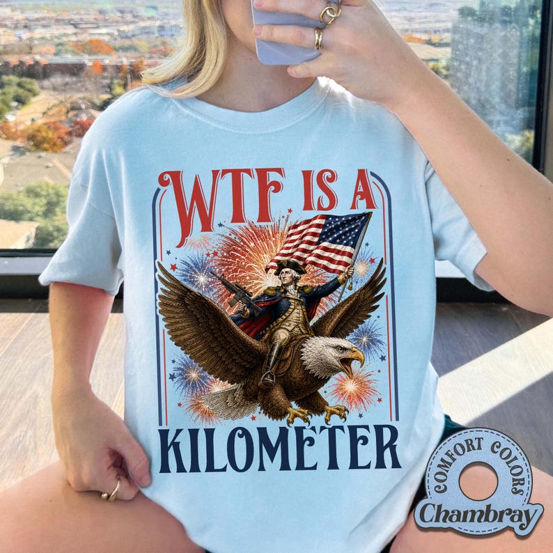 May include: Light blue t-shirt with a patriotic graphic. The design features an eagle carrying a figure holding a flag, with fireworks and the text "WTF IS A KILOMETER". The shirt has a round neckline and short sleeves.