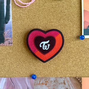 May include: A heart-shaped embroidered patch featuring a dark brown center with a white stylized TW logo. The patch has a layered design with a bright orange section on the right and a deep pink section on the left, all bordered by a dark brown edge.