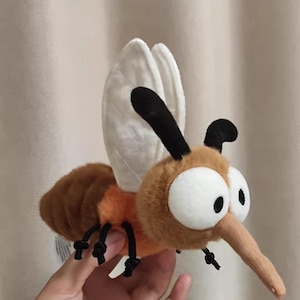 Op de afbeelding: Een pluche muggenspeelgoed met grote witte ogen, een lange beige snuit en een bruin en oranje lichaam. Het speelgoed heeft witte vleugels en zwarte poten en antennes. De mug is gemaakt van zacht, pluizig materiaal.