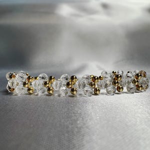 Op de afbeelding: Een heldere kristallen armband met goudkleurige kralen. De armband is gemaakt van kleine, facetgeslepen kristallen en afgewisseld met goudkleurige, bolvormige kralen. De sieraden worden gepresenteerd op een zilveren satijnen oppervlak.