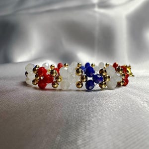 Op de afbeelding: Een armband met rode, witte, blauwe en gouden kralen. De armband is gemaakt met kleine, ronde kralen in een bloemmotief. De kralen zijn geregen met een goudkleurige draad. De armband ligt op een witte achtergrond.
