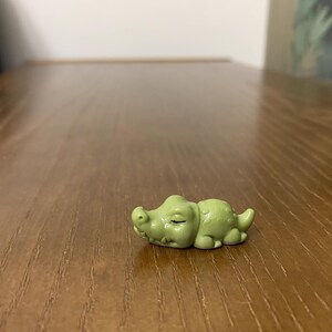 Könnte beinhalten: Kleine, hellgrüne Alligatorfigur in Ruheposition. Die Figur hat geschlossene Augen und liegt auf einer Holzoberfläche. Der Alligator hat eine hellgrüne Farbe.