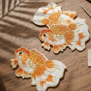 May include: Two embroidered patches shaped like goldfish, featuring intricate stitching in shades of orange, gold, and white. Each patch depicts a fish with a rounded body, prominent fins, and a detailed tail, showcasing textured thread work.