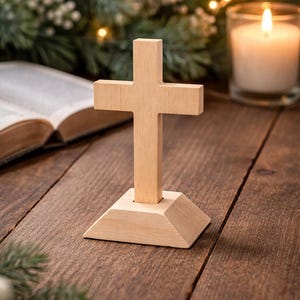 Puede incluir: Una cruz de madera clara sobre un soporte, con una vela encendida y un libro abierto en el fondo. La cruz está sobre una base geométrica. El fondo presenta vegetación y una iluminación suave, creando una escena serena.