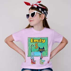 May include: A light pink t-shirt featuring a cartoon cat with the name "Emily" and the number "5" on a green chalkboard. The cat wears a blue shirt, jeans, and mismatched boots. The person in the image is wearing heart-shaped sunglasses and a red, white, and blue headband.