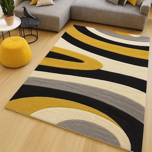 May include: A rectangular area rug with a modern design. The rug features curved stripes in black, mustard yellow, cream, and gray. The rug is placed on a light wood floor, with a gray sectional sofa and yellow accents in the background.