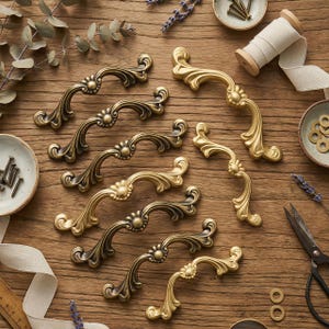 Peut inclure: Diverses poignées de meubles décoratives en laiton antique et dorées. Les poignées présentent un design incurvé avec des détails floraux. Divers accessoires de quincaillerie, dont des vis et du ruban, sont disposés sur une surface en bois.