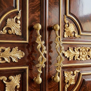 Puede incluir: Primer plano de una puerta de armario de madera con adornos y tiradores de hojas de oro ornamentados. La puerta es de un marrón oscuro intenso con un acabado brillante. Los tiradores son dorados, con un diseño de hojas y vides.