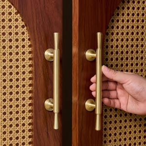 May include: Close-up of a wooden cabinet with brass handles. The cabinet doors are a rich, dark brown, and the handles are vertical with a cylindrical design. The background features a woven cane pattern.