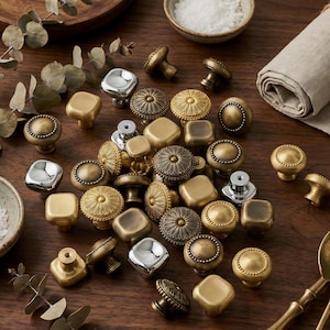May include: An assortment of decorative cabinet knobs in various shapes and finishes, including silver, gold, and bronze. The knobs feature detailed designs, such as floral patterns and rounded edges. The hardware is arranged on a wooden surface.