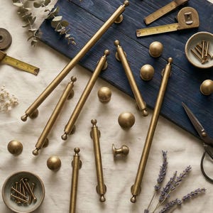 May include: A collection of antique brass cabinet hardware, including pulls, knobs, and a handle, displayed on a blue wooden surface. A measuring tape, small bowls with screws, and scissors are also present. The hardware has a warm, golden colour.