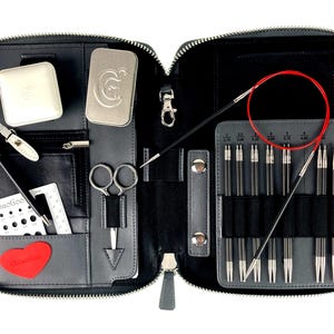 May include: A black leather case with a zipper closure, containing a set of interchangeable knitting needles, a pair of scissors, a measuring tape, a needle gauge, and a small tin with a logo. The case is open, revealing the contents.