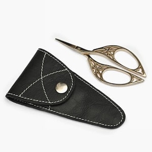 May include: A pair of gold-colored embroidery scissors with ornate handles rests beside a black leather sheath. The sheath is triangular, with white stitching and a silver snap closure. The scissors are approximately 4 inches long.