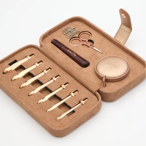 May include: A brown leather case with a gold zipper and a gold clasp. The case contains a set of knitting needles, a pair of scissors, a tape measure, and a small wooden tool. The case is open and the contents are visible.