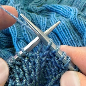 May include: Close-up of knitting needles with blue yarn creating a textured fabric. The needles are silver, and the yarn has shades of blue and teal. The image shows the process of knitting, with stitches in progress.