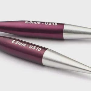 May include: Two knitting needles with a plum-colored body and silver tips. The text "6.0mm - US10" is printed on the body of each needle. The needles are lying on a light gray surface.