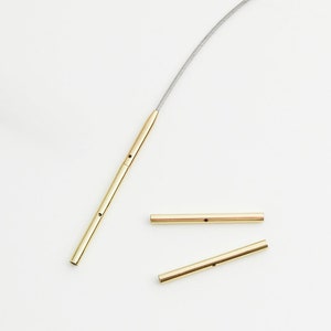 May include: A close-up of several gold-colored metal components. One piece is attached to a thin, silver-colored wire. Two additional pieces are lying separately. Each piece has small holes.