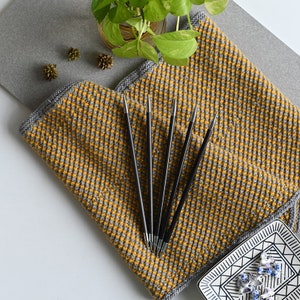May include: A set of black knitting needles with silver tips, lying on top of a yellow and gray knitted fabric. The fabric has a checkered pattern.
