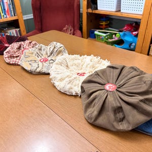 May include: A collection of six round, flat caps arranged in a row. The caps feature various fabrics including dark red velvet, red and white plaid, a beige paisley print, a cream-colored fringed texture, solid brown cotton, and blue denim.