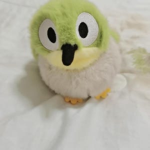 May include: A plush toy bird with a green head, white body, and yellow feet. The bird has large, round eyes with black pupils and a black beak. The toy is made of soft, fluffy material and is sitting on a white surface.