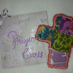 May include: A handmade prayer cross made of colorful fabric patches with an orange stitched border. A matching card with the words "Prayer Cross" in purple script and a decorative floral pattern is included.