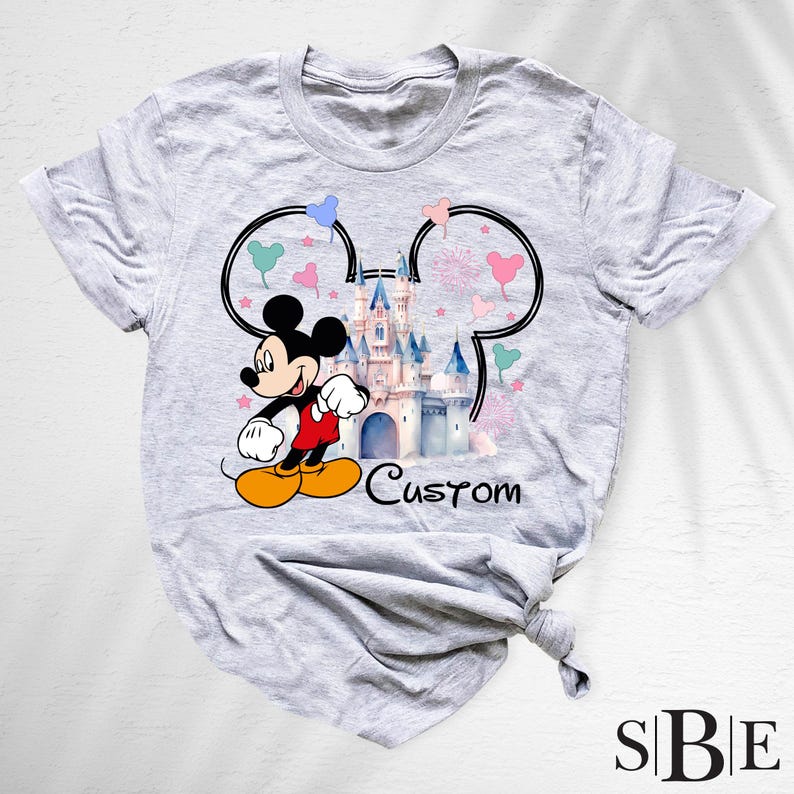 May include: A light gray t-shirt featuring a colorful graphic of Mickey Mouse in front of a castle. The design includes Mickey Mouse ears, hearts, and the word "Custom". The shirt has short sleeves and a crew neck.