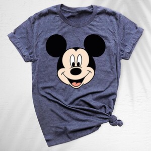 May include: A blue t-shirt featuring a large graphic of Mickey Mouse's face. The cartoon character's face is black, white, and tan, with a smiling expression. The shirt has short sleeves and a crew neck.
