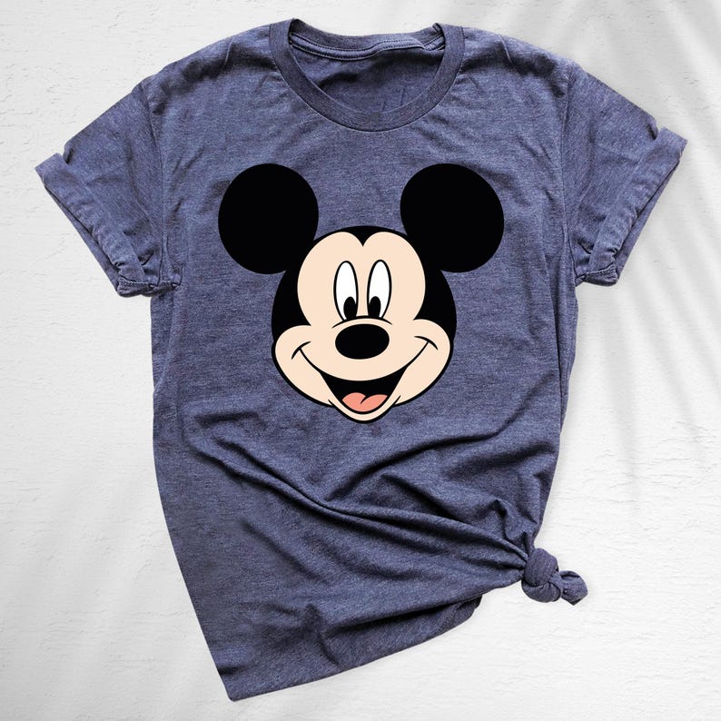 May include: A blue t-shirt featuring a large graphic of Mickey Mouse's face. The cartoon character's face is black, white, and tan, with a smiling expression. The shirt has short sleeves and a crew neck.
