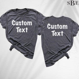 May include: Two dark heather gray short-sleeve t-shirts with crew necklines. Each shirt features the white text Custom Text printed on the front. The fabric has a visible heathered texture, and both shirts are knotted at the bottom hem.