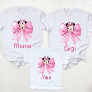 以下が含まれることがあります： ミニーマウスのデザインが施された白いTシャツ3枚。各シャツにはピンクのリボンとミニーの顔が描かれています。「ママ」、「ジジ」、「ミニ」とピンクの文字で書かれており、家族でお揃いに最適です。
