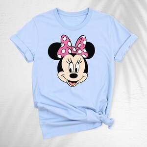 May include: A light blue t-shirt featuring a graphic of Minnie Mouse's face. Minnie's face is black with a pink bow with white polka dots. The t-shirt has rolled-up sleeves and a knot at the bottom.