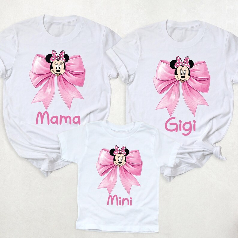 以下が含まれることがあります： ミニーマウスのデザインが施された白いTシャツ3枚。各シャツにはピンクのリボンとミニーの顔が描かれています。「ママ」、「ジジ」、「ミニ」とピンクの文字で書かれており、家族でお揃いに最適です。
