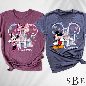 May include: Two t-shirts, one maroon and one blue, each featuring a Disney castle design with Mickey and Minnie Mouse. The word "Custom" is printed below the castle. The shirts have a crew neck and short sleeves.