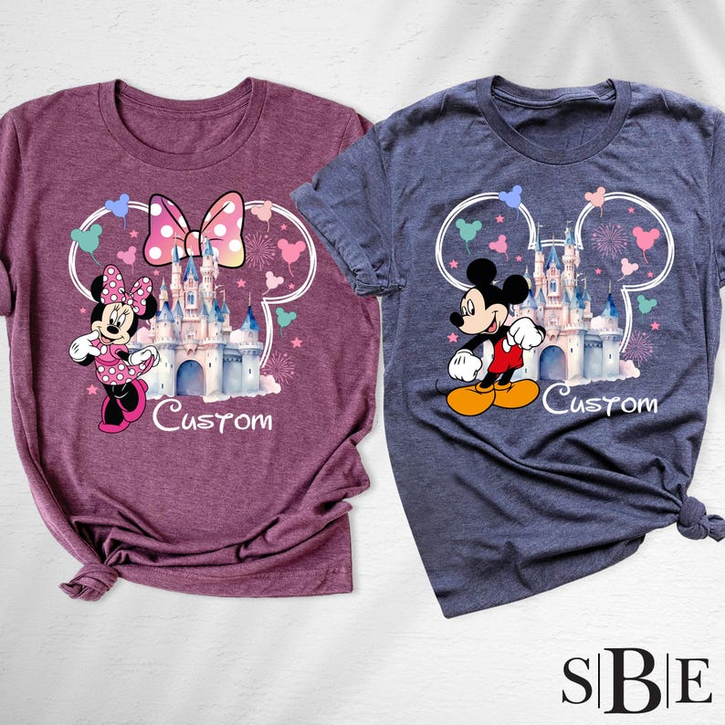 May include: Two t-shirts, one maroon and one blue, each featuring a Disney castle design with Mickey and Minnie Mouse. The word "Custom" is printed below the castle. The shirts have a crew neck and short sleeves.