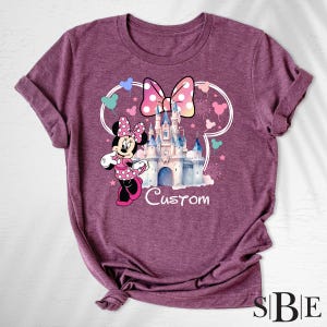 May include: Burgundy t-shirt featuring a Minnie Mouse graphic. The design includes Minnie Mouse, a castle, and a polka dot bow. The word "Custom" is printed below the castle. The shirt has rolled-up sleeves.