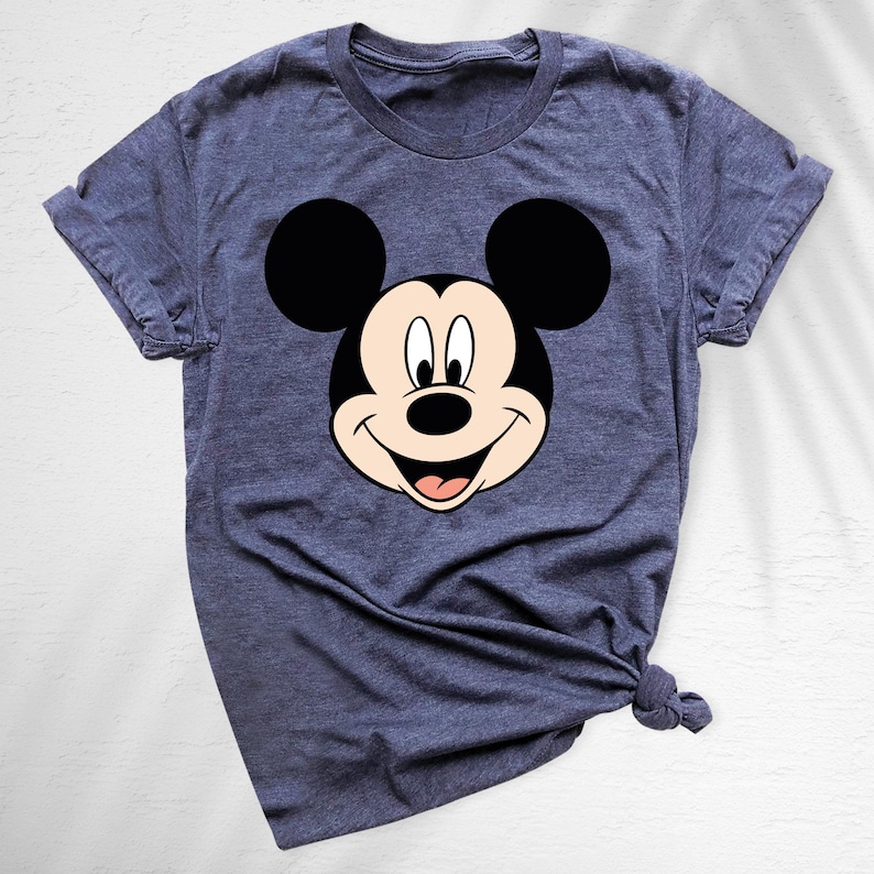 May include: A blue t-shirt featuring a large graphic of Mickey Mouse's face. The cartoon character's face is in black, white, and tan, with a smiling expression. The shirt has short sleeves and a crew neck.