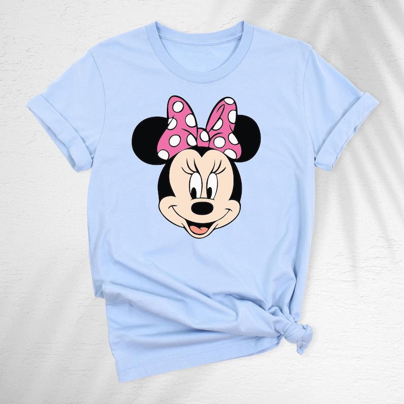May include: A light blue t-shirt featuring a graphic of Minnie Mouse's face. Minnie's face is black with a pink bow with white polka dots. The t-shirt has rolled-up sleeves and a knot at the bottom.