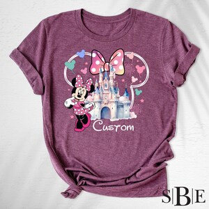May include: Burgundy t-shirt featuring a Minnie Mouse graphic with a polka dot bow, castle, and the word "Custom". The design is framed by a white outline of Mickey Mouse ears and colorful accents.
