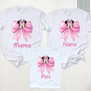 以下が含まれることがあります： ミニーマウスのデザインが施された白いTシャツ3枚。各シャツにはピンクのリボンとミニーの顔が描かれています。シャツにはピンクの文字で「ママ」、「ナナ」、「ミニ」と書かれています。シャツは半袖です。