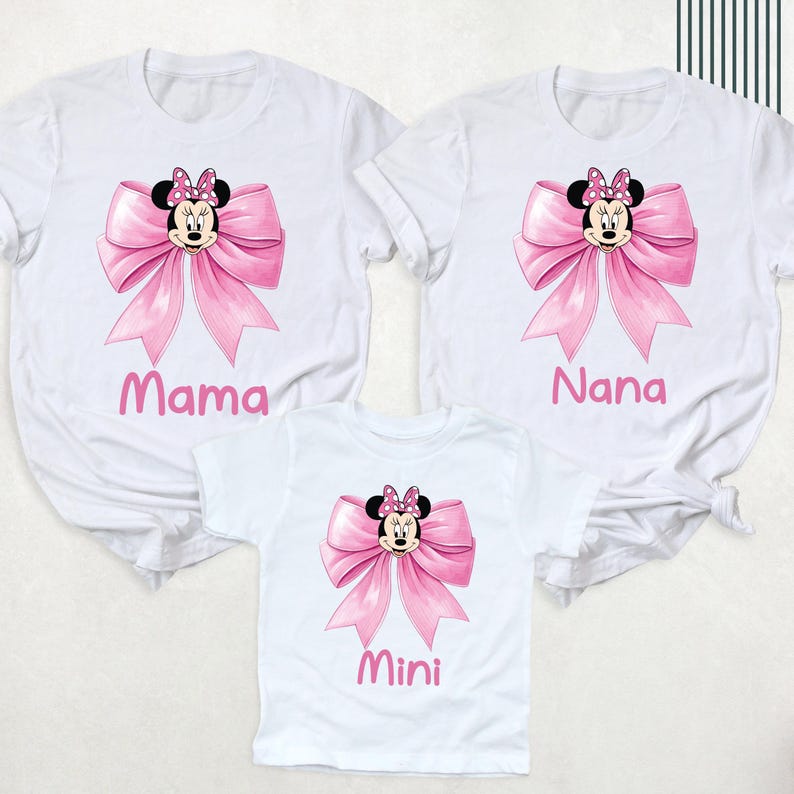 以下が含まれることがあります： ミニーマウスのデザインが施された白いTシャツ3枚。各シャツにはピンクのリボンとミニーの顔が描かれています。シャツにはピンクの文字で「ママ」、「ナナ」、「ミニ」と書かれています。シャツは半袖です。