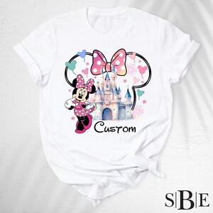 May include: White t-shirt featuring a Minnie Mouse graphic with a pink polka dot bow, a castle, and the word "Custom". The design includes a Minnie Mouse character and heart shapes.