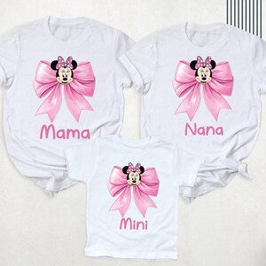 以下が含まれることがあります： ミニーマウスのデザインが施された白いTシャツ3枚。各シャツにはピンクのリボンとミニーの顔が描かれています。シャツにはピンクの文字で「ママ」、「ナナ」、「ミニ」と書かれています。シャツは半袖です。