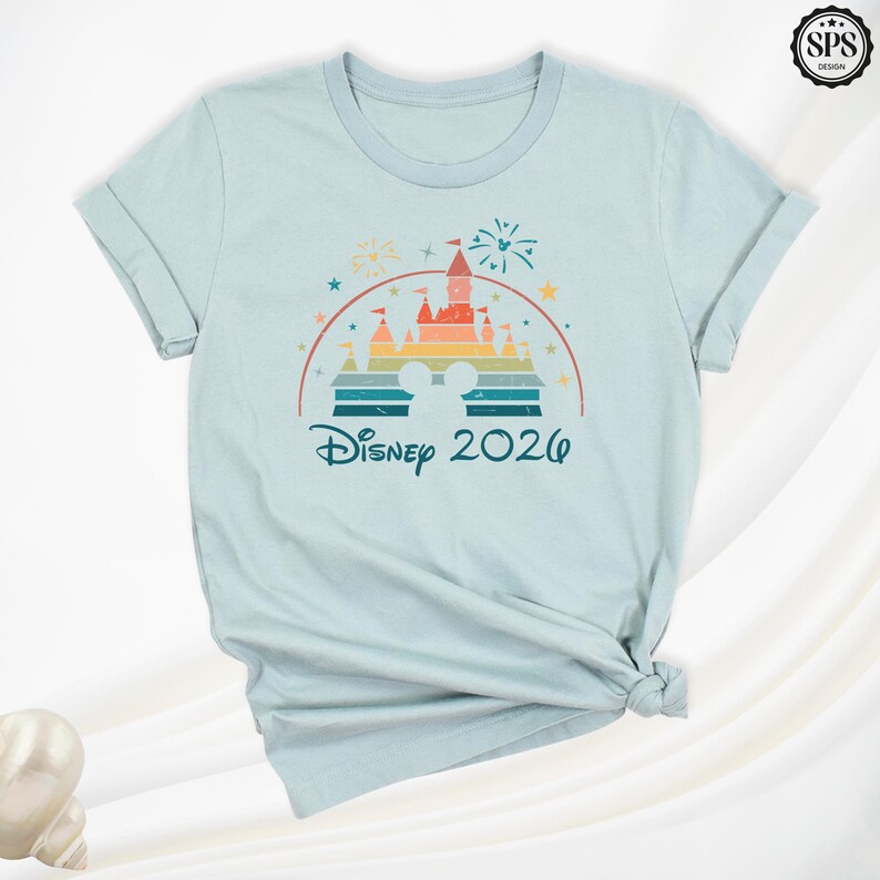 May include: Light blue t-shirt featuring a colorful graphic of a castle, fireworks, and the text "Disney 2026". The design includes a rainbow arch and Mickey Mouse ears. The shirt is made of a soft material.