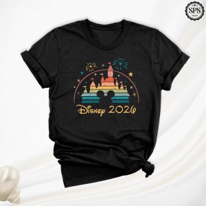 May include: Black t-shirt featuring a colorful graphic of a castle, fireworks, and Mickey Mouse ears. The text "Disney 2026" is printed below the graphic in a vintage style. The shirt has short sleeves and a crew neck.