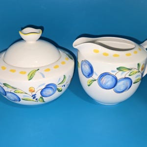 May include: A white ceramic sugar bowl with a lid and a creamer jug. Both items are decorated with blue plums, green leaves, and yellow accents. The sugar bowl has a yellow-trimmed knob on the lid.