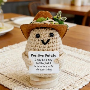 Könnte beinhalten: Eine gehäkelte Kartoffel-Figur mit einem lächelnden Gesicht, die einen braunen Hut mit grünen Blättern und weißen Akzenten trägt. Die Kartoffel hält ein Schild mit der Aufschrift "Positive Potato" und einer inspirierenden Botschaft. Sie ist auf einem Holzsockel montiert.