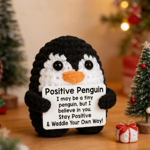 Peut inclure: Un pingouin crocheté noir et blanc avec un bec orange et des yeux noirs. Il tient une pancarte avec l'inscription "Positive Penguin" et un message motivant. L'arrière-plan comprend de petits arbres de Noël et des décorations festives.