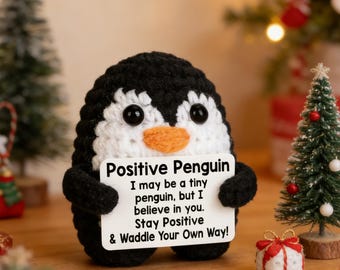 Pode incluir: Um pinguim de crochê em preto e branco com bico laranja e olhos pretos. Ele segura uma placa com a inscrição "Positive Penguin" e uma mensagem motivacional. O fundo inclui pequenas árvores de Natal e decorações festivas.
